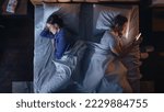 Small photo of Top View Bedroom Apartment: Young Woman Uses Smartphone in Bed at Night When Her Male Partner Trying to Fall Asleep Beside. Couple Fight, Argue. Social Media, Doom Scrolling, Fake News Addiction