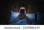 Small photo of Anonymous Man Uses Smartphone in Bed at Home at Night. Handsome Guy Browsing Social Media, Reading News, Doing Online Shopping Late at Night. Focus on Hand Holding Mobile Phone Covering Face
