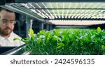 Small photo of Portrait of an Agricultural Grower Working in a Corridor in a Vertical Farm Facility Next to a Rack with Freshly Grown Plants. Caucasian Hydroponics Technician Closely Studying and Cultivating Crops