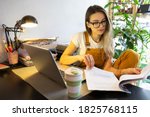 Small photo of Millennial woman gardener in yellow overalls using laptop, examines project documentation, makes a report on paper documents, works from home. Cozy office workplace, remote work, distant education.