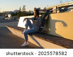 Young girl use smartphone sitting outdoors at urban park with longboard texting with friends via mobile phone messenger application online. Casual teen female with cellphone chatting at sunset outside