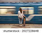 Young girl passenger with longboard standing on subway station platform with blurry moving blue train on background, rear view. Woman with skateboard watching metro pass fast at the station.