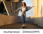 Young carefree woman enjoy riding skateboard at sunset. Happy girl in trendy street style clothes, hipster eyeglasses and jeans skating longboard over urban background. Freedom and lifestyle concept