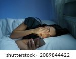 Tired woman lying in bed sleepless holding smartphone in hand reading about sleep disorder and mental health in dark bedroom. Anxious female need rest. Sleeplessness, anxiety and depression concept
