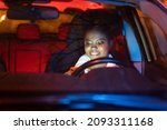 Small photo of African woman distracted on smart phone while driving car at night. Smiling black female read text on mobile phone sitting on driver seat. Irresponsible girl messaging riding vehicle. Safety on road.