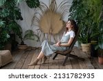 Small photo of Mental health benefits of gardening. Young happy female gardener sitting in chair surrounded by various lush tropical plants. Smiling woman resting in home garden. Botanic boom and urban jungle trend