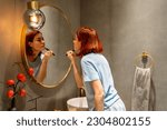 Small photo of Redhead girl looking in mirror applying everyday makeup on face with brush. Young woman standing in bathroom using compact powder to cover flaws. Fitting in society beauty standards