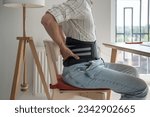 Small photo of Man freelancer works types on computer keyboard wearing back support belt corset on lower back during exacerbation to treatment of hernia. Back pain health problems as consequences of sedentary work.