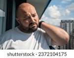 Small photo of Bearded man in hot summer sun on balcony. Bald, unsmiling guy closed eyes in displeasure discontent from discomfort, holding on to sore neck, kneading ligaments, suffering from protrusion, pain, ache