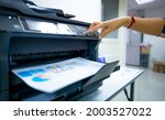 Small photo of Office worker prints paper on multifunction laser printer. Copy, print, scan, and fax machine in office. Document and paper work. Print technology. Hand press on photocopy machine. Scanner equipment.