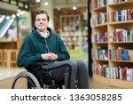 Content handsome young disabled student with headphones on neck siting in wheelchair and looking at camera in modern library or bookstore