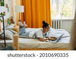 Full length portrait of carefree teenage girl using laptop on bed in cozy room, copy space