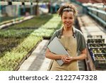 Pretty young African American female farmer or woner oh large hothouse with laptop standing against long tables with seedlings