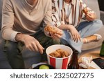 Small photo of Closeup young couple eating fried chicken takeout while watching TV at home, copy space