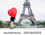 Man holding a woman on his arms (lifting a woman) and she is hiding their faces with the heart shape red umbrella. Trocadero square with the view to the city Paris and Eiffel Tower
