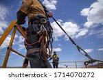 Small photo of Safe workplace rigger wearing working at heights harness clipping an inertia reel shock absorbing fall arrest device hook on the rope sling anchor point while working from 2 metre exposure open edges