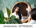 Small photo of Hair care and self-care with beautiful Indian woman looking in mirror touching her healthy long hair sitting at the dressing table.