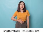 Small photo of Pleased cheerful Asian woman keeps hand on belly feels full after delicious dinner dressed casually stands thoughtful against blue background.