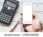 Hand holding smartphone white display with silver pen on blank page diary calculator and gold coins group on white wood table top view nature shadow,Business concept