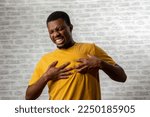 Small photo of Young man presses hand to chest has heart attack suffers from unbearable pain closes eyes poses against grey brick wall background. People young age and problems with health concept, copy space.