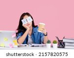 Small photo of Sleep deprivation. Young asian business woman sitting at workplace with stickers on eyes and holding cup with coffee, working on laptop at table pastel pink background