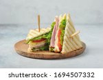 Classic club sandwich on a wooden plate. The sandwich filling includes ham, cheese, lettuce, tomato slices. Light background. Close-up.