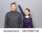 Small photo of Studio shot of short woman standing on tiptoes and showing height of tall man, both are cheerful and smiling, over gray background. Concept of diversity of people's heights, tall and short persons