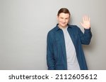 Small photo of Studio portrait of happy mature man raising hand, waving palm, saying hello, smiling joyfully, looking friendly and amiable, wearing blue shirt, standing over gray background, with copy space