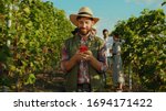 Small photo of Joyful bearded farmer wearing strawhat browsing fun content on smartphone during rest on grapevine alley at vineyard. Wine worker. People and technology.