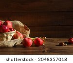 Basket with red apples on sackcloth close up. Dry oak leaves, acorns, ripe fruits on wooden table at brown wood barn wall background. Autumn food, fall menu, thanksgiving, harvest season concept