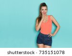 Beautiful young woman in orange tank top is holding hand on chin and talking. Three quarter length studio shot on turquoise background.