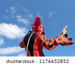 Small photo of Person in a jester costume isolated on blue sky with white clouds, carnival concept. Harlequin in black-red foolscap with bells, girl dressed as a Joker