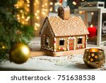 Little gingerbread house with glaze standing on table with tablecloth and decorations, candles and lanterns. Living room with lights and Christmas tree. Holiday mood