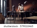 Handsome serious tailor in white shirt with brown leather suspenders working near wooden table with threads, apron and scissors in amazing atelier with antique furniture and mannequin on background