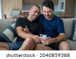 Small photo of couple of gay men in love, relaxing on the sofa in their living room, fiddling with their cell phone, researching the new summer vacation