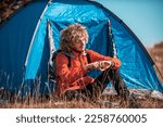 Small photo of A man camping with a tent. He is surrounded by a beautiful mountain landscape, and the sun shines on his face, foreshadowing a beautiful and sunny day. The man looks satisfied and fulfilled