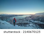 Female Adventurer in a red jacket and backpack stands atop a fjellheisen and watches Norwegian Sea surrounding the city of Tromso in northern Norway above the Arctic Circle on frosty winter morning.