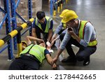 Small photo of Safety colleagues team helping middle aged warehouse asian worker who had broken head accident and lying on the floor in warehouse while using walkie talkie radio. First aid training concept.