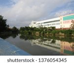 Small photo of Surabaya, Indonesia - April 21 2019: View of the back of JMP Grocery Mall Market Building with clear blue sky clouds and river background with trees around. Nearby with Red Bridge Historical Duct area