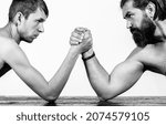 Small photo of Heavily muscled bearded man arm wrestling a puny weak man. Arms wrestling thin hand, big strong arm in studio. Black and white.