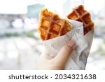 Small photo of Food Trend - Croffles (compound word from Croissant and Waffle) wrapped with food grade oil absorbing paper holding by female hand on blurred cityscape background. (selective focus, space for text)