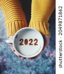 Small photo of Number 2022 on frothy surface of cappuccino served in white cup holding by female hands over rustic blue background. Holidays food art theme Happy New Year 2022, New year new you. (selective focus)