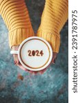 Small photo of Happy New Year 2024 theme number 2024 on frothy surface of cappuccino served in white coffee mug holding by female hands over rustic blue background. Holidays food art, new year new you. (top view)