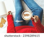 Small photo of 2024 New Year's goal setting, number 2024 on frothy surface of cappuccino in white coffee cup holding by woman in red knitted sweater with jeans sitting on bed while writing down her resolutions.