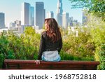  A young brunette woman sits with her back to the camera on a park bench overlooking Downtown Los Angeles skyscrapers