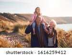 Grandfather Giving Granddaughter Ride On Shoulders As They Walk Through Sand Dunes With Grandmother