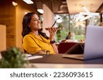 Small photo of Mature Businesswoman Working On Laptop At Desk In Office Talking Into Mic Of Mobile Phone