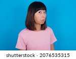 Small photo of young asian woman wearing pink t-shirt against blue background with snobbish expression curving lips and raising eyebrows, looking with doubtful and skeptical expression, suspect and doubt.