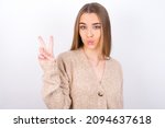 young caucasian girl wearing knitted sweater over white background makes peace gesture keeps lips folded shows v sign. Body language concept