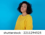 Small photo of brunette arab woman wearing yellow sweater over blue background with snobbish expression curving lips and raising eyebrows, looking with doubtful and skeptical expression, suspect and doubt.